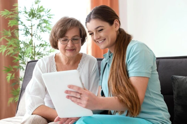 A private caregiver and a older woman, are seated on a couch, engaged with a tablet between them.