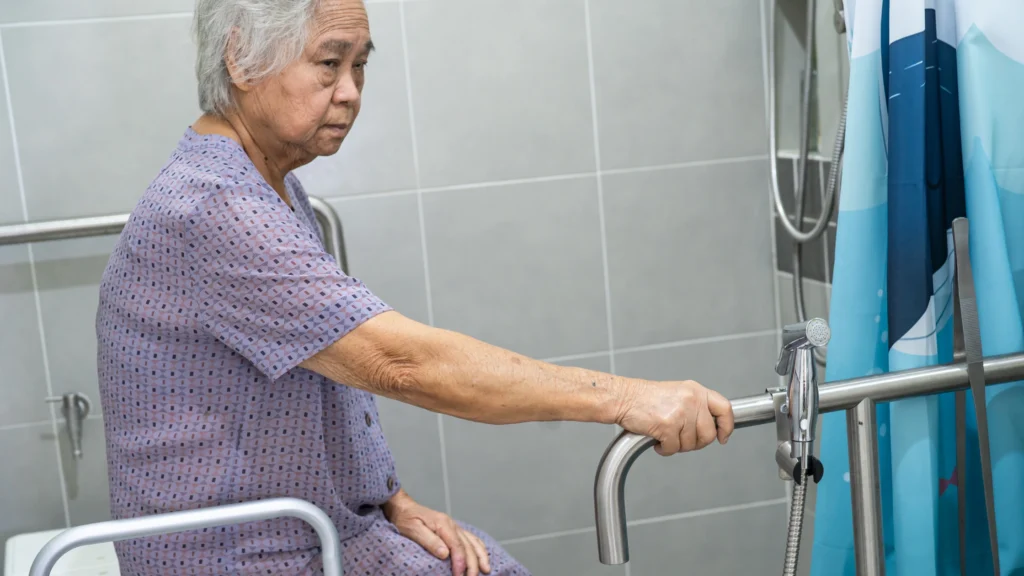 Elderly woman in a bathroom gripping a safety rail for support. Proper bathroom modifications help ensure safety when bathing seniors at home.