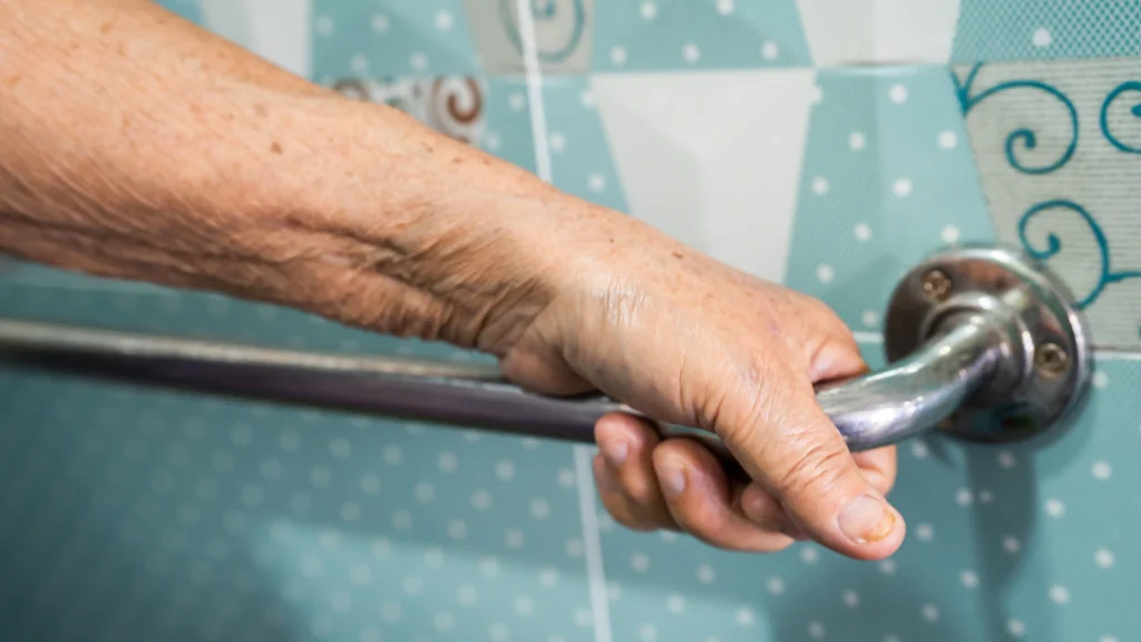 Senior hand gripping a wall-mounted grab bar in a bathroom. Using sturdy handrails can significantly reduce fall risks when assisting elderly individuals with bathing at home.