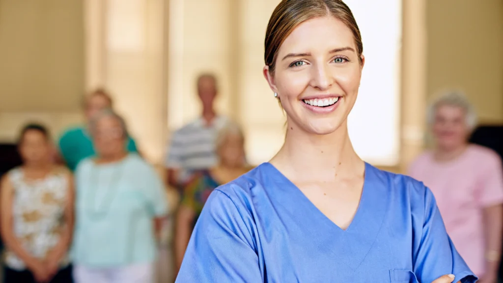 A smiling independent caregiver in blue scrubs stands confidently in front of a group of elderly individuals, symbolizing professional and compassionate senior care.