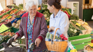 An independent caregiver helps an elderly woman with a walker while grocery shopping at an outdoor market, showcasing personalized support and companionship.