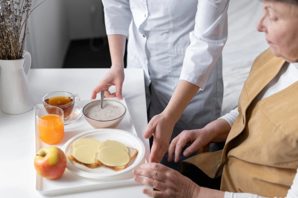 A caregiver serving a healthy breakfast with cheese toast, porridge, tea, juice, and an apple to an elderly woman, ensuring proper nutrition.