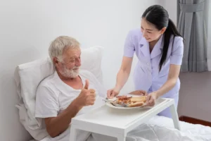 A smiling nurse serves breakfast to an elderly man in bed, who gives a thumbs-up, illustrating compassionate at-home senior care.