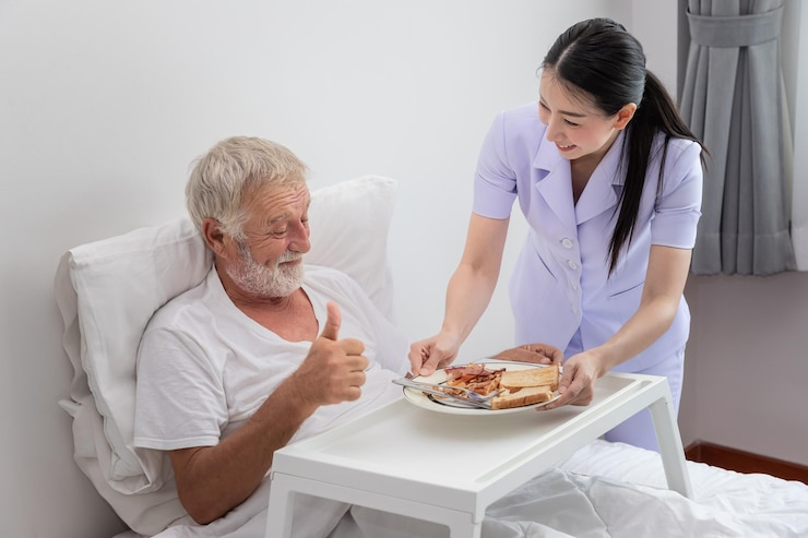 A smiling nurse serves breakfast to an elderly man in bed, who gives a thumbs-up, illustrating compassionate at-home senior care.