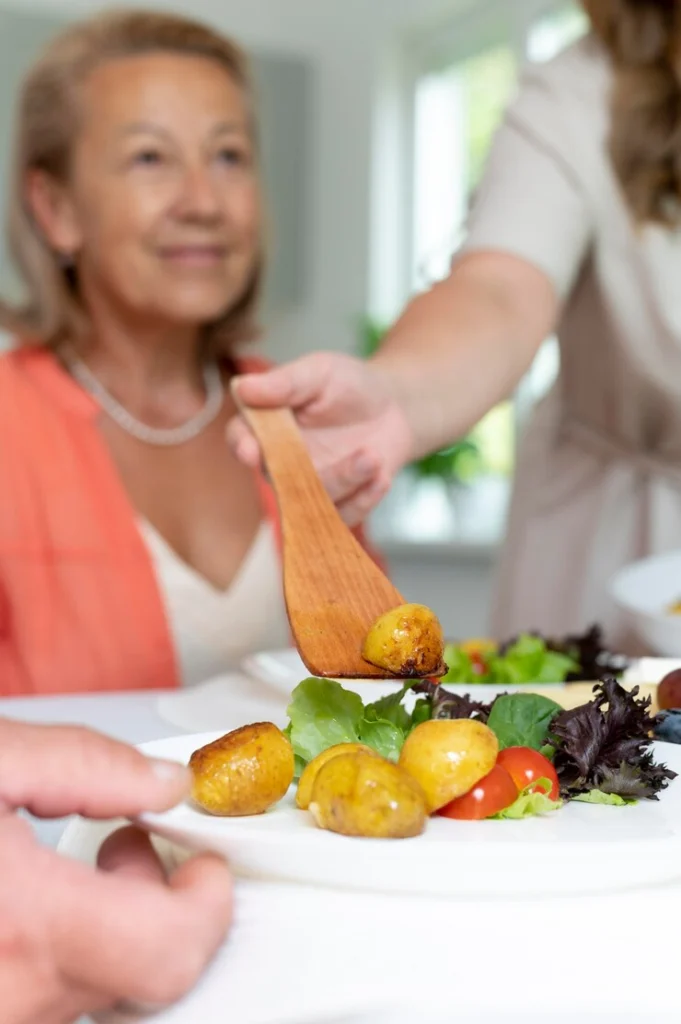 A caregiver serving a nutritious meal with roasted potatoes, fresh salad, and cherry tomatoes to an elderly woman, promoting healthy meals for elderly.